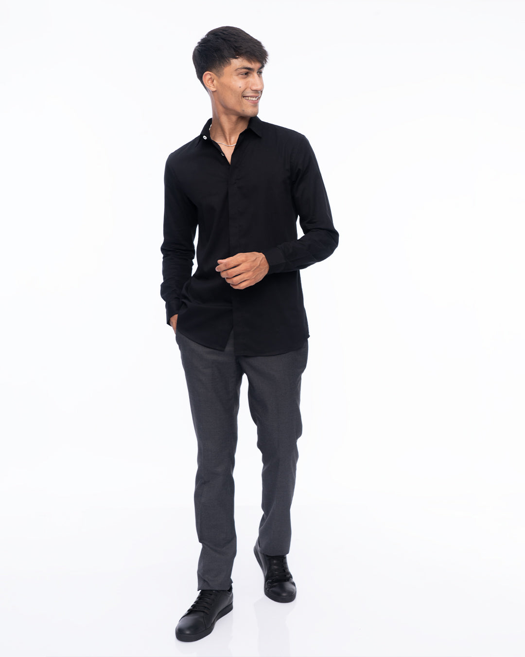 A person with short dark hair wearing an Obsidian - Classic Shirt made from premium cotton and gray pants is smiling while standing with their right hand in their pocket against a plain white background.