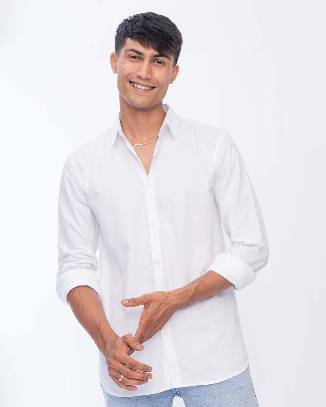 A person with short dark hair is smiling at the camera, wearing a Crystal - Classic Shirt with the sleeves slightly rolled up. One arm is relaxed by their side, and the other hand is resting on it. The background is plain white.