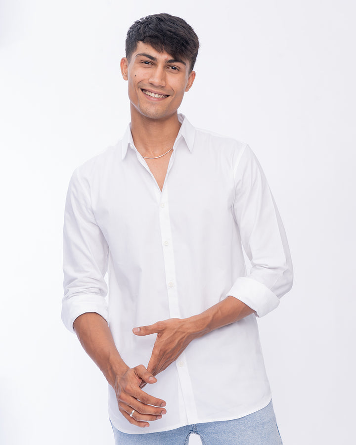 A person with short dark hair is smiling at the camera, wearing a Crystal - Classic Shirt with the sleeves slightly rolled up. One arm is relaxed by their side, and the other hand is resting on it. The background is plain white.