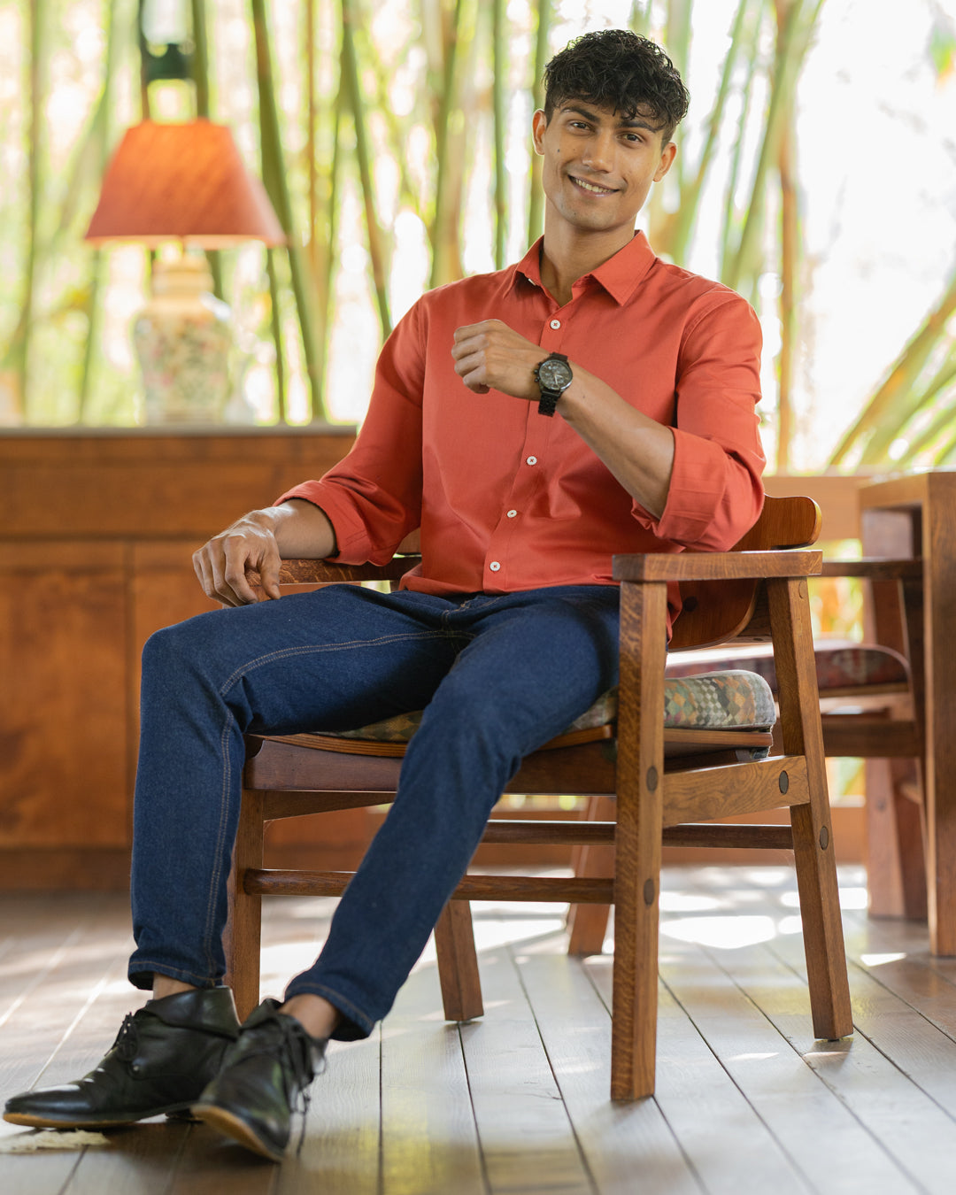 A man with dark hair, wearing a red shirt crafted from premium cotton and blue jeans, is sitting on a wooden chair indoors. He is smiling and has one arm rested on the chair's armrest while the other hand is slightly raised. The background features wooden furniture and soft lighting from the Scarlatto - Classic Shirt.