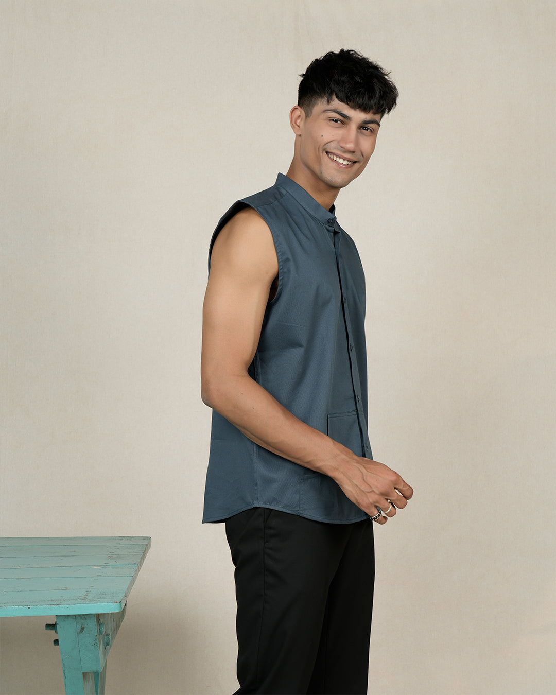 A smiling man with short, dark hair stands against a plain background. He is wearing a sleeveless, button-up, dark gray Sarcelle - Sleeveless Shirt made of premium cotton with a mandarin collar and black pants. He has a wristwatch on his left hand and stands near a small, light blue table.