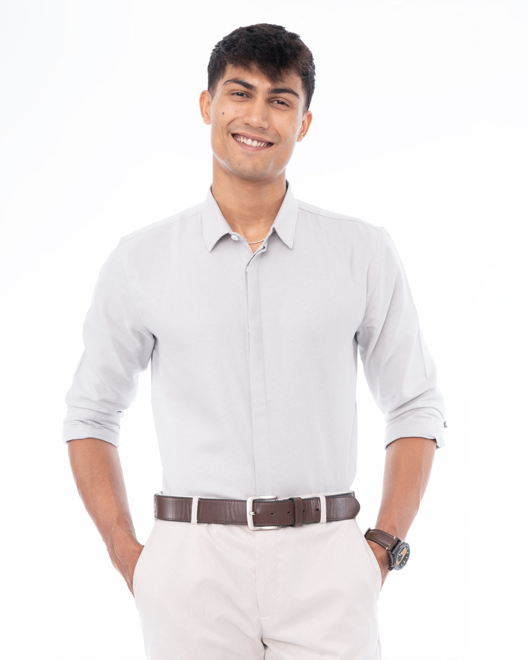 A man with short dark hair smiles at the camera. He is wearing a light gray, long-sleeved, Ash Grey - Classic Shirt and white pants. With one hand in his pocket, he stands against a plain white background, showcasing perfect everyday wear.