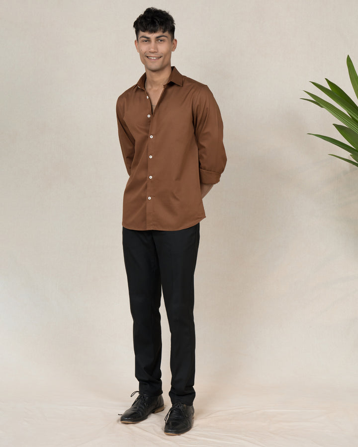 A smiling person with short, dark hair stands against a neutral background, wearing a casual Brunn - Classic Shirt with rolled-up sleeves and black pants. They have one hand in their pocket and the other slightly extended forward. A green plant is partially visible on the right.