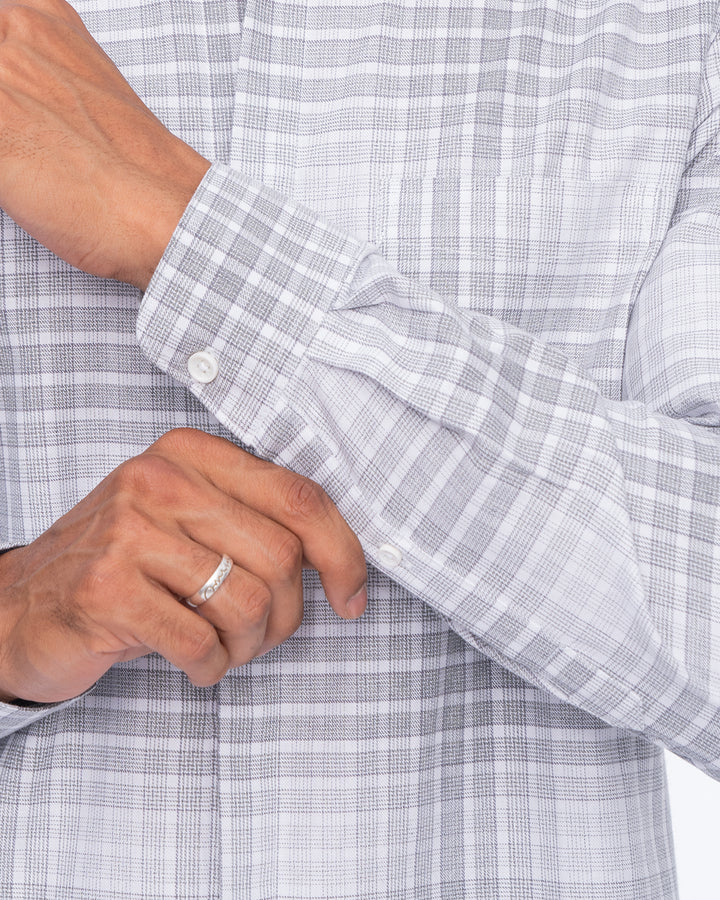 A man wearing a light grey, checkered Pebble - Mandarin Shirt made of premium cotton with rolled-up sleeves is standing against a plain white background. He is smiling and has his hands clasped in front of him.