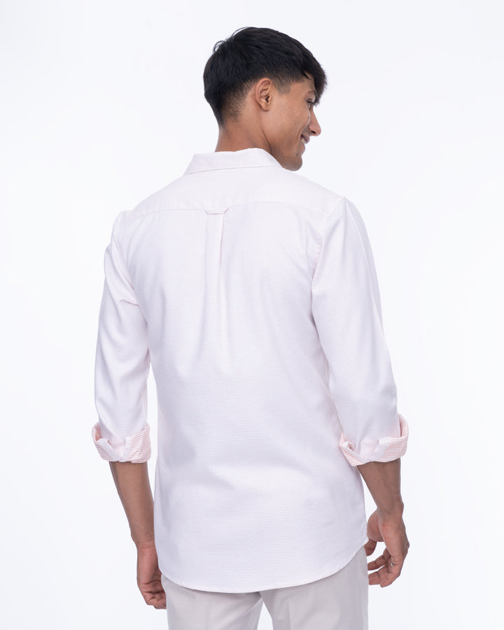 A smiling man stands against a white background, wearing a light pink cotton shirt with the sleeves rolled up and light gray pants. This Ivory - Classic Shirt is perfect for everyday wear as he looks to his right, with his hands relaxed in front of him.