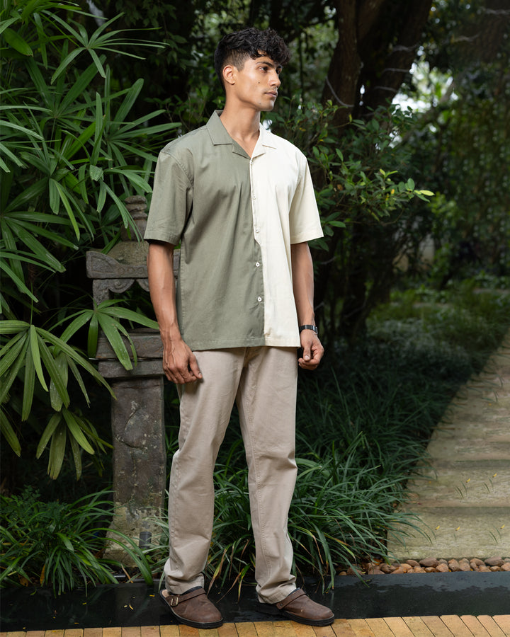 A man with curly dark hair smiles while standing in front of greenery. He is wearing a short-sleeved Dark Sage Moitie - Cuban Collar Shirt that is olive green on one side and cream on the other, paired with light-colored pants. He stands with one hand in his pocket.