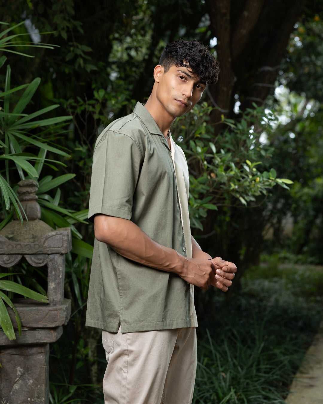 A man with curly dark hair smiles while standing in front of greenery. He is wearing a short-sleeved Dark Sage Moitie - Cuban Collar Shirt that is olive green on one side and cream on the other, paired with light-colored pants. He stands with one hand in his pocket.