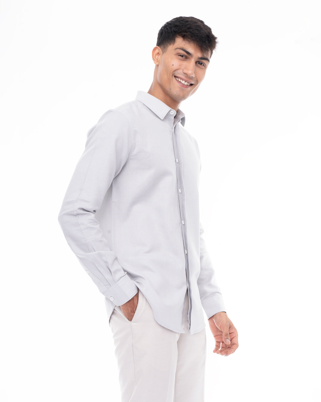 A man with short dark hair smiles at the camera. He is wearing a light gray, long-sleeved, Ash Grey - Classic Shirt and white pants. With one hand in his pocket, he stands against a plain white background, showcasing perfect everyday wear.