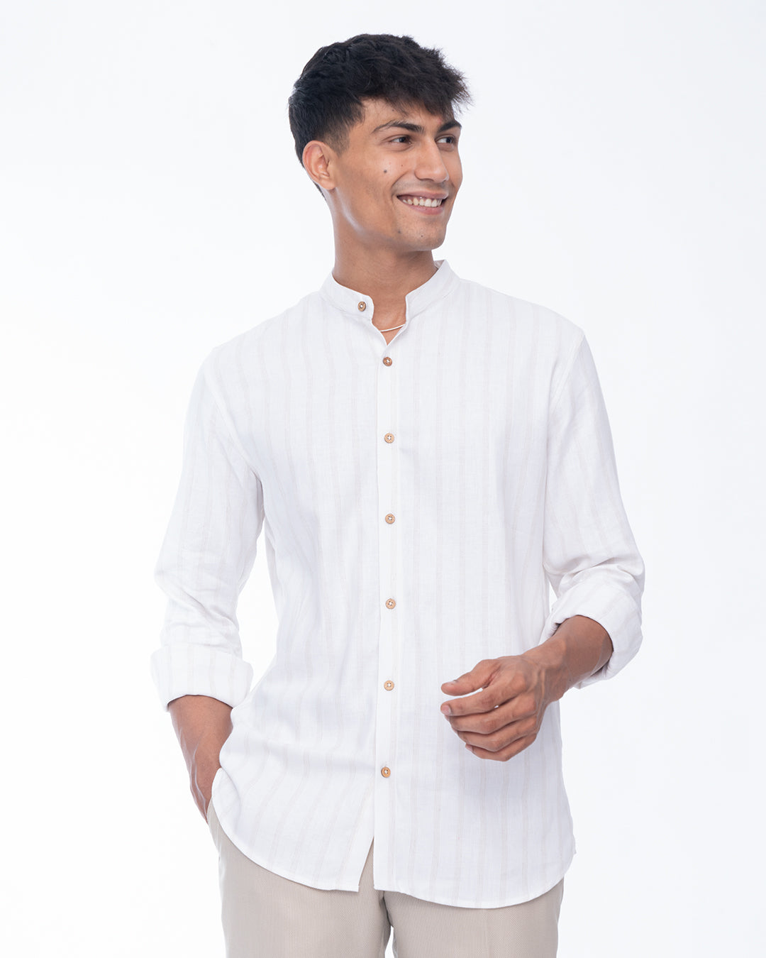 A person with short dark hair is smiling and wearing a Seashell White - Mandarin Shirt made from premium cotton. They have one hand in their pocket and the other hand is holding the edge of their shirt. The background is a plain white studio setting.