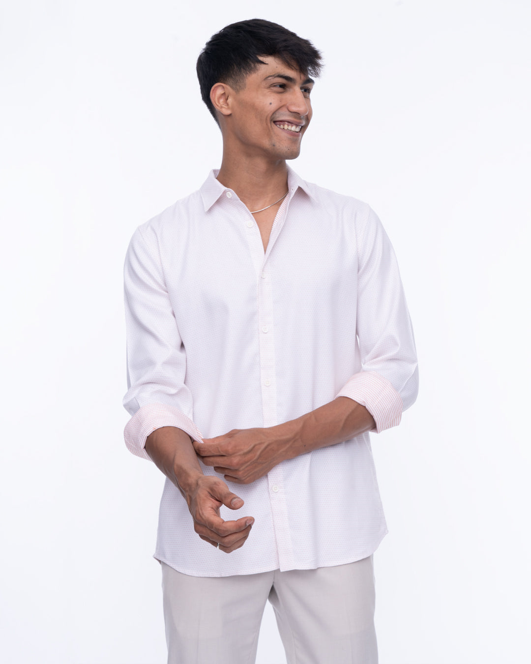 A smiling man stands against a white background, wearing a light pink cotton shirt with the sleeves rolled up and light gray pants. This Ivory - Classic Shirt is perfect for everyday wear as he looks to his right, with his hands relaxed in front of him.