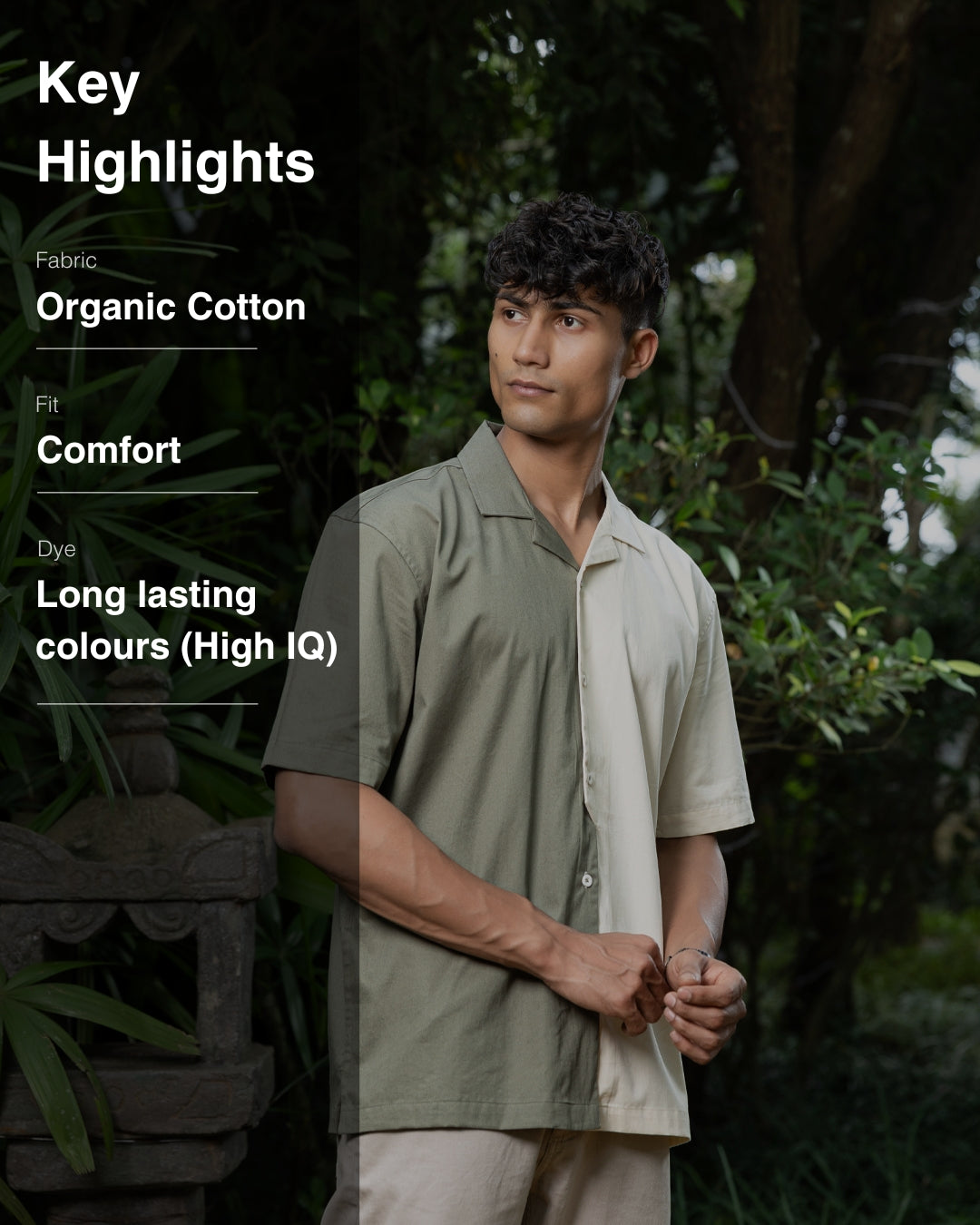 A man with curly dark hair smiles while standing in front of greenery. He is wearing a short-sleeved Dark Sage Moitie - Cuban Collar Shirt that is olive green on one side and cream on the other, paired with light-colored pants. He stands with one hand in his pocket.