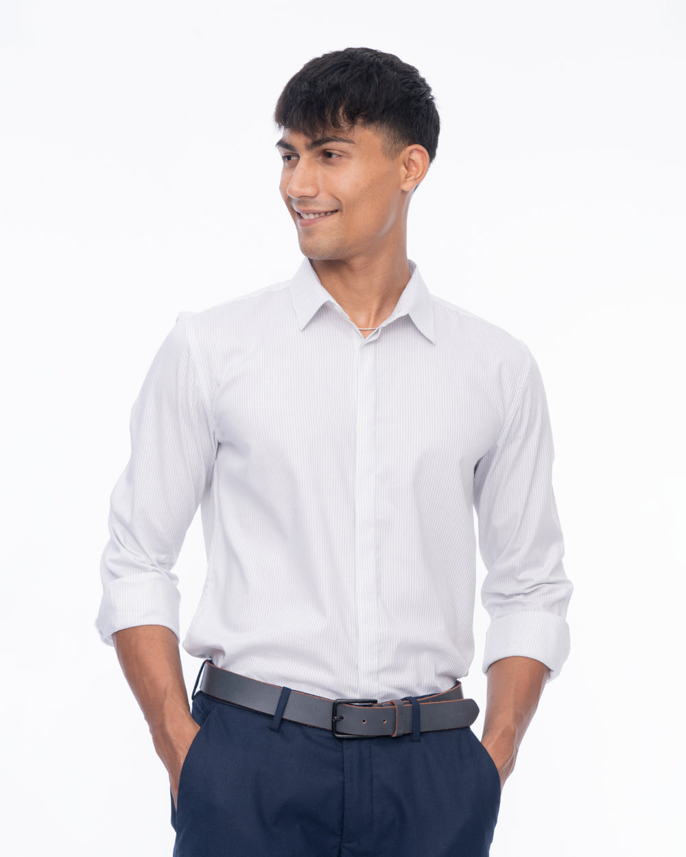 A young man stands against a plain white background, smiling and wearing a Sky Line - Classic Shirt made from premium cotton paired with dark pants. His hands are relaxed and resting in his pockets.