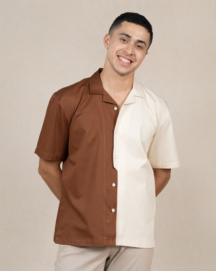 A smiling man wearing a short-sleeved Brunn Moitie - Cuban Collar Shirt from the High IQ Dye Collection. The unique design features a vertical split, with one half in rich brown and the other in light cream, made from premium cotton. He stands against a neutral background with his hands behind his back.