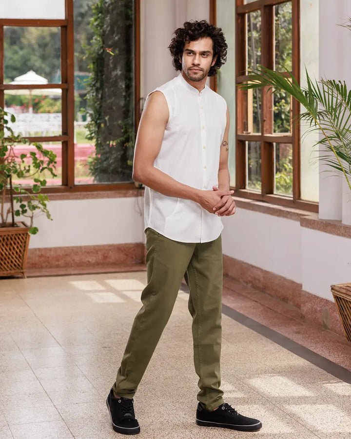 A man with curly hair is standing indoors, wearing a Branco - Sleeveless Shirt made of premium cotton and green pants. He has a tattoo on his left arm and is smiling slightly. Behind him, there are large windows with lush greenery visible outside.